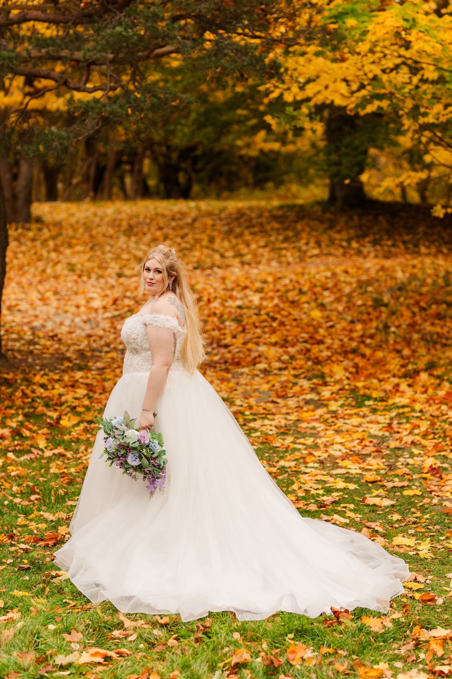 A flowing white wedding dress spreads across vibrant autumn leaves in a golden forest setting.