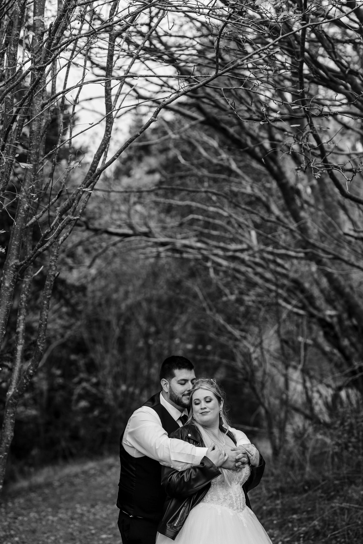 A romantic black and white wedding portrait captured in a wooded setting with intertwined branches overhead.