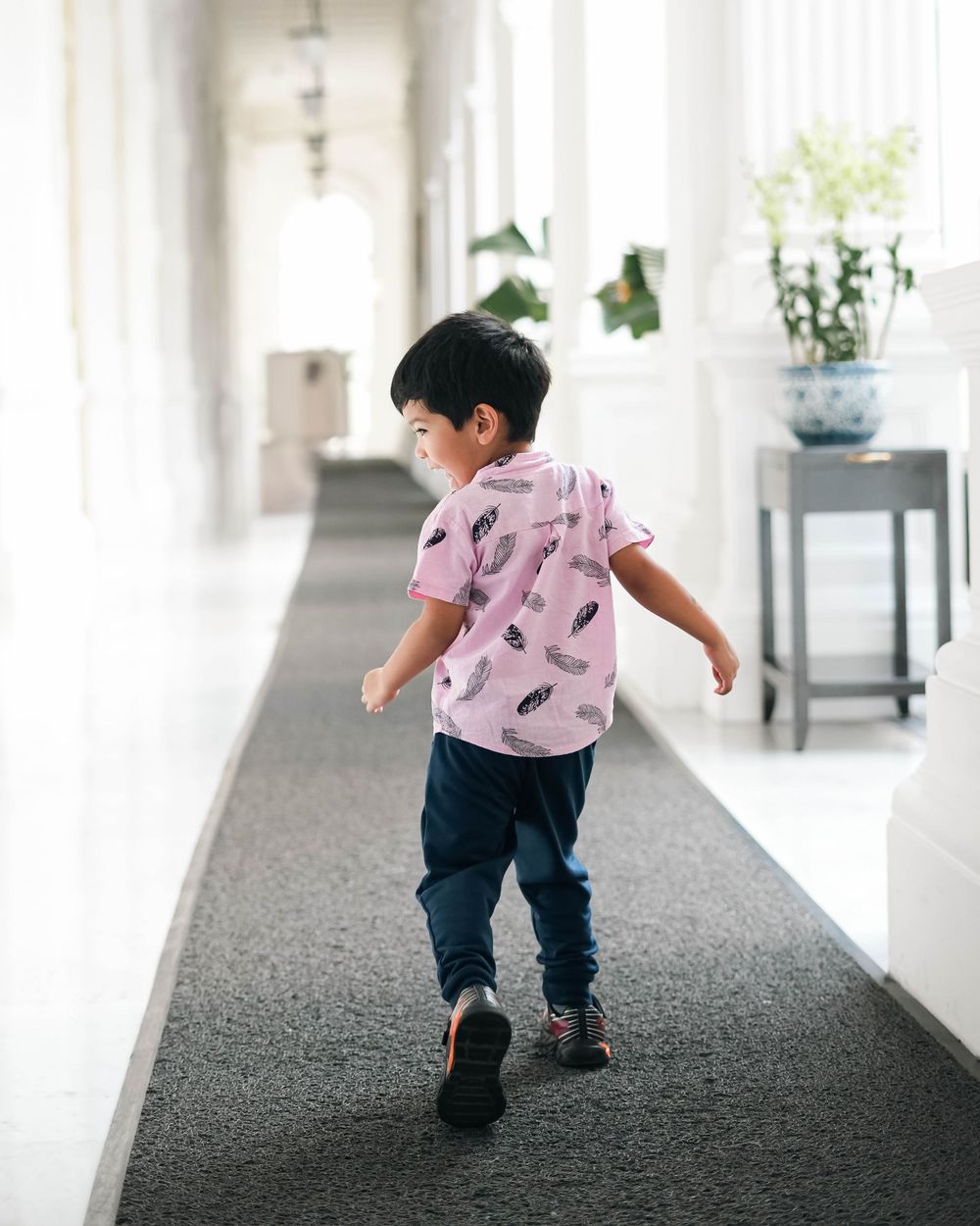 A toddler in a pink shirt and jeans runs playfully down a carpeted hallway with natural light streaming in.