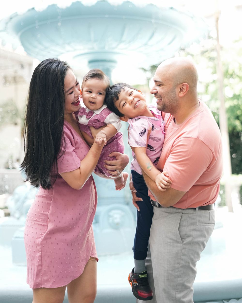 A joyful family shares a candid moment of laughter together by a blue fountain in matching pink and peach attire.