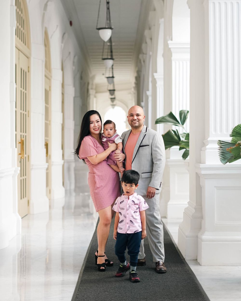A family poses together in a white colonnade hallway with elegant architectural details and pendant lighting.