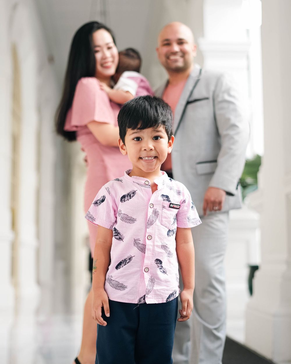 A family in coordinated pink and gray outfits smiles together during a photo session in a bright hallway.