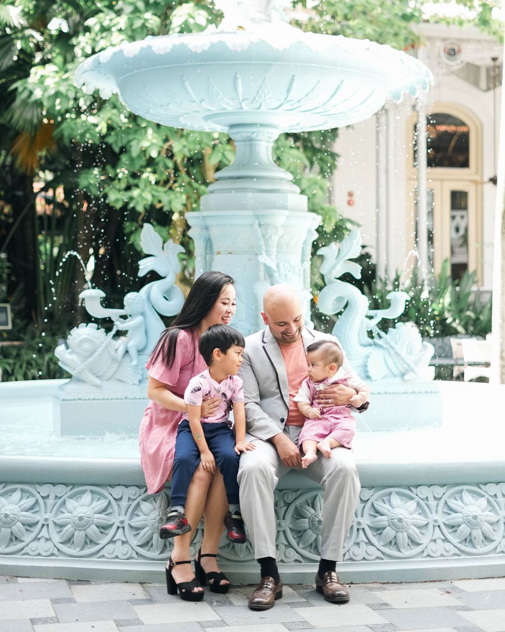 People sit together on an ornate white stone fountain ledge in a garden setting with lush greenery.