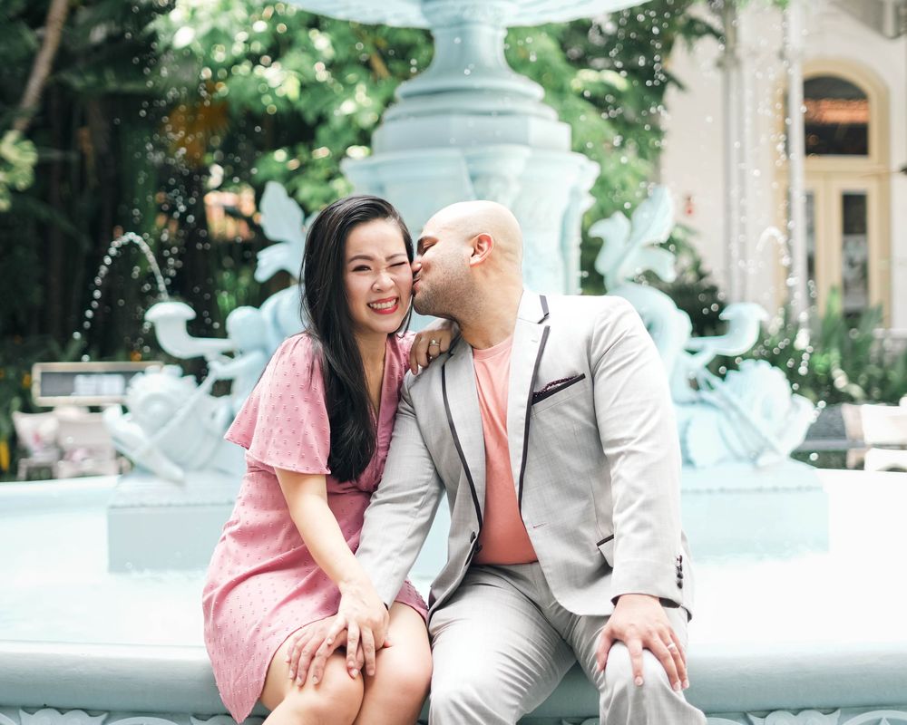 A couple shares a romantic moment together on an ornate white stone bench in a garden setting.