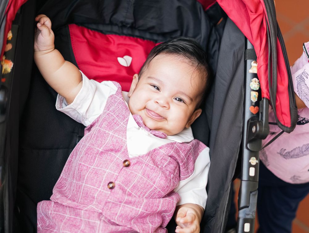 A baby wearing a pink plaid shirt smiles and waves while sitting in a high chair.