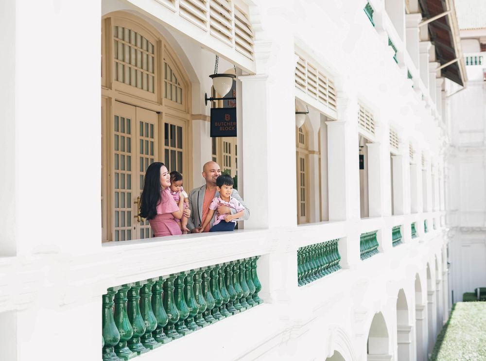 Two people in pink outfits stand by white colonial-style pillars and green railings in a bright corridor.