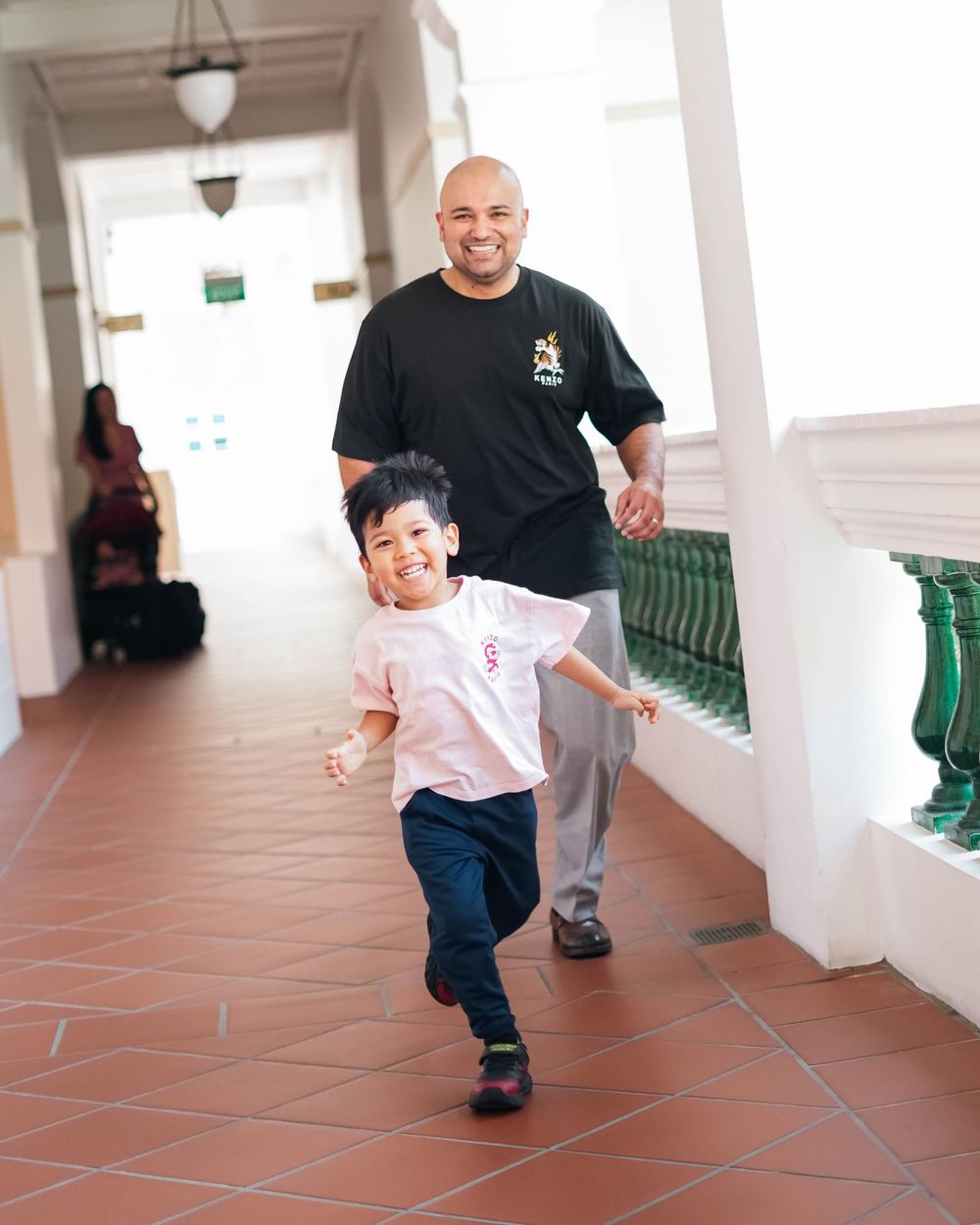 A joyful child runs playfully down a covered outdoor corridor with terracotta flooring while an adult follows behind.