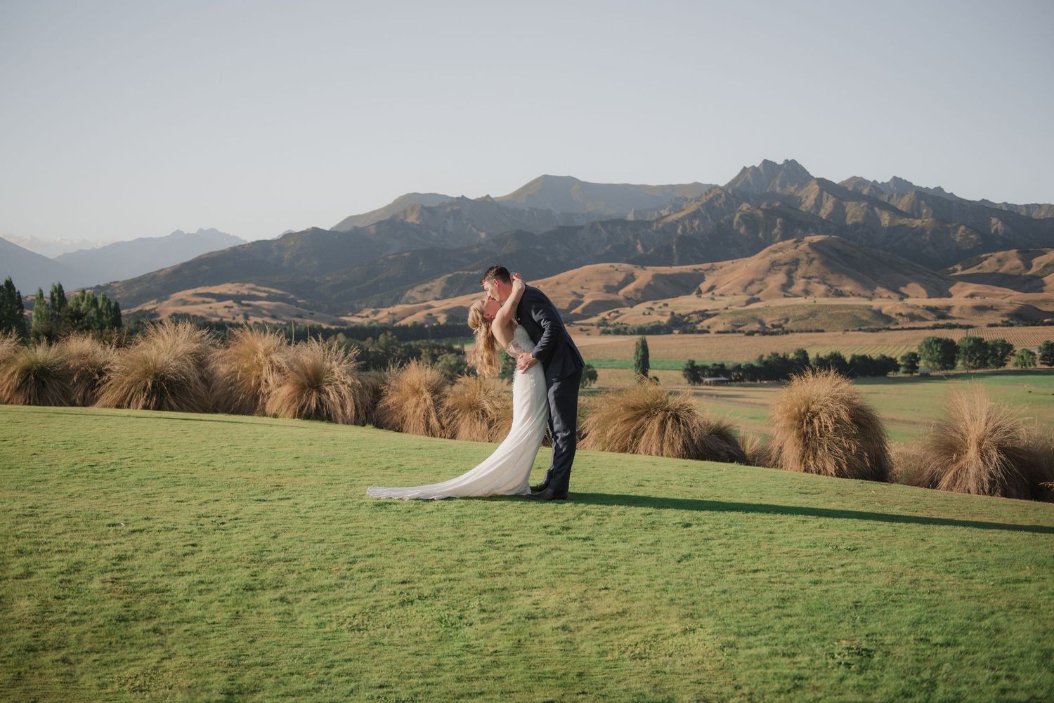A couple embraces on a hilltop golf course with majestic mountains in the background during golden hour.