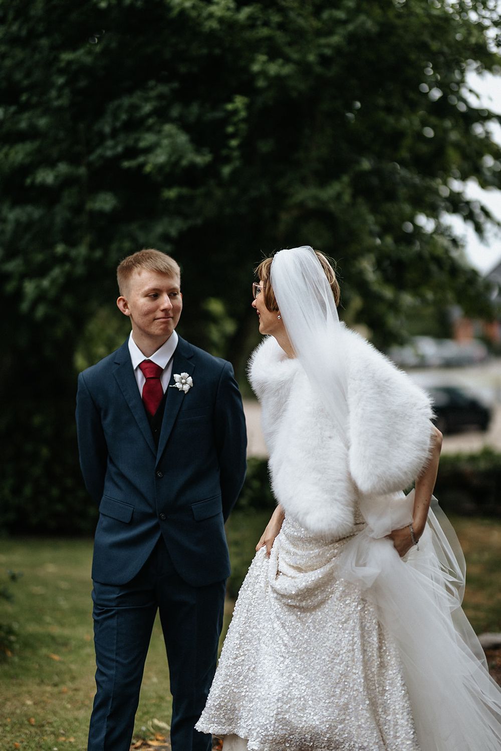 Wedding couple poses together outdoors with trees in background, bride wearing white fur shawl.