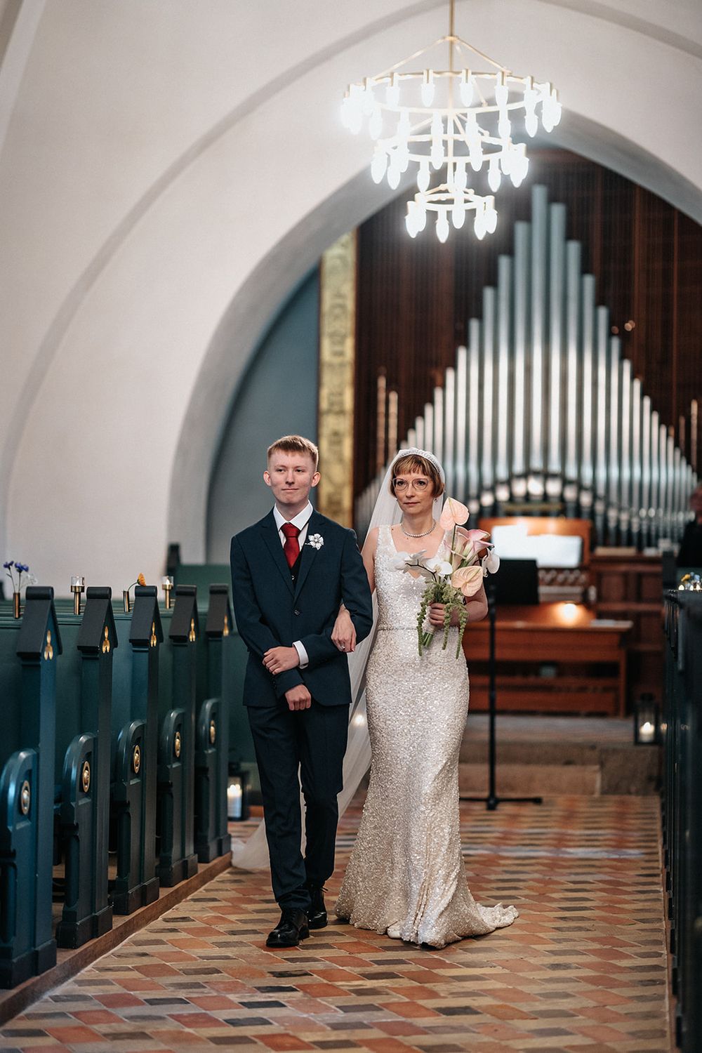 Wedding ceremony processional in church aisle with ornate chandeliers and organ pipes visible.