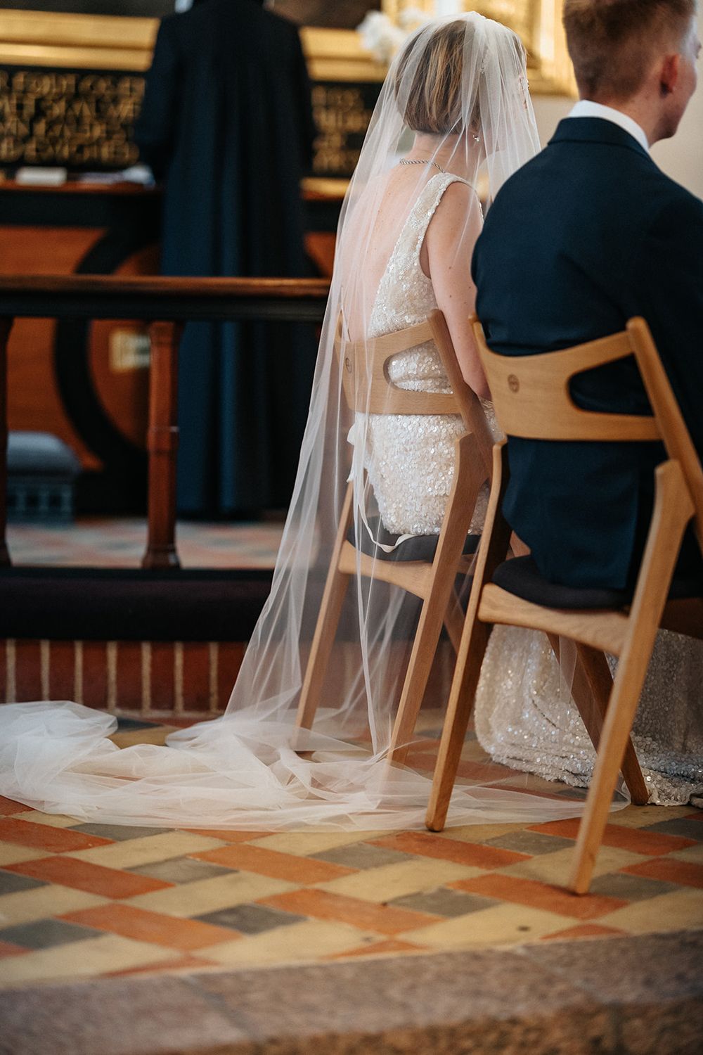 A bride in a white lace dress with flowing veil sits on a wooden chair on a patterned tile floor during a wedding ceremony.