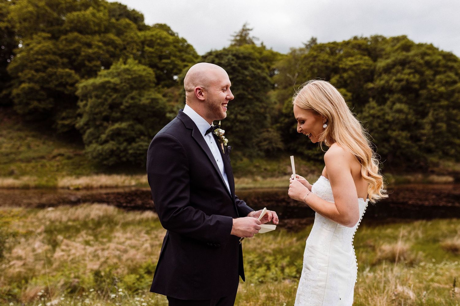 A couple stands in a meadow of white wildflowers surrounded by lush green trees under a cloudy summer sky.