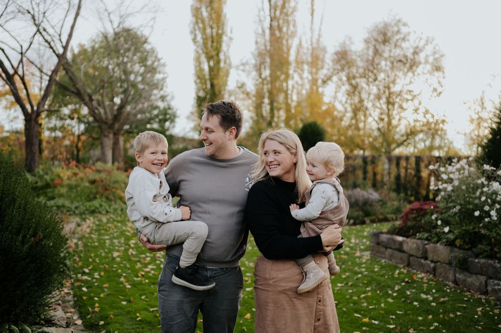 Eine vierköpfige Familie genießt gemeinsam fröhliche Momente auf einer Wiese mit herbstlichen Bäumen im Hintergrund.