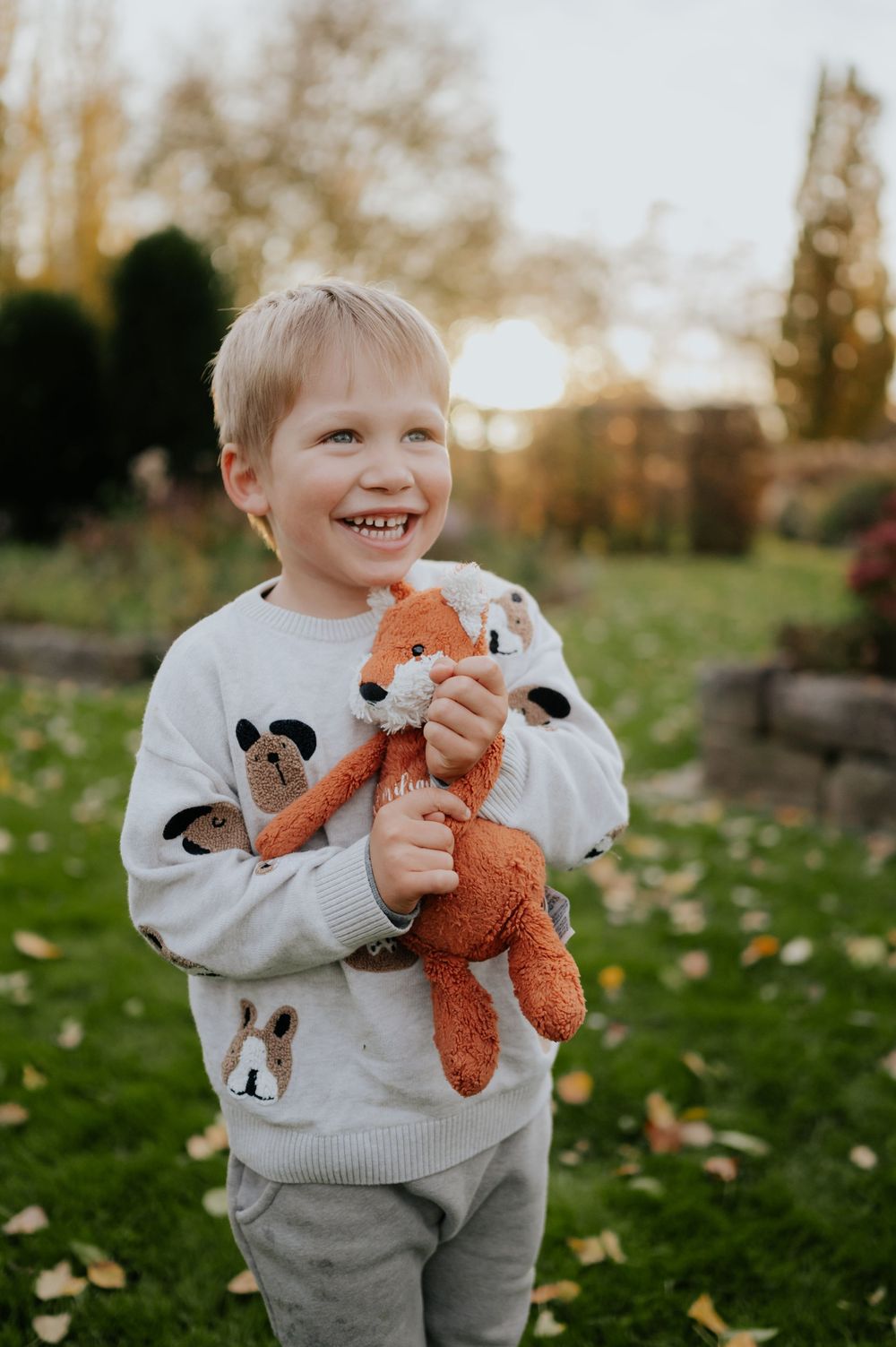 Kind in einem Pullover mit Panda-Aufdruck hält ein orangefarbenes Stofftier in Form eines Fuchses in der Hand und lächelt draußen bei Sonnen