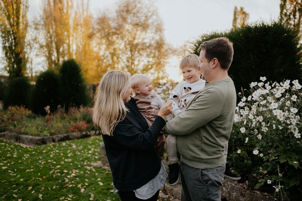 Eine vierköpfige Familie genießt gemeinsam fröhliche Momente auf einer Wiese mit herbstlichen Bäumen im Hintergrund.