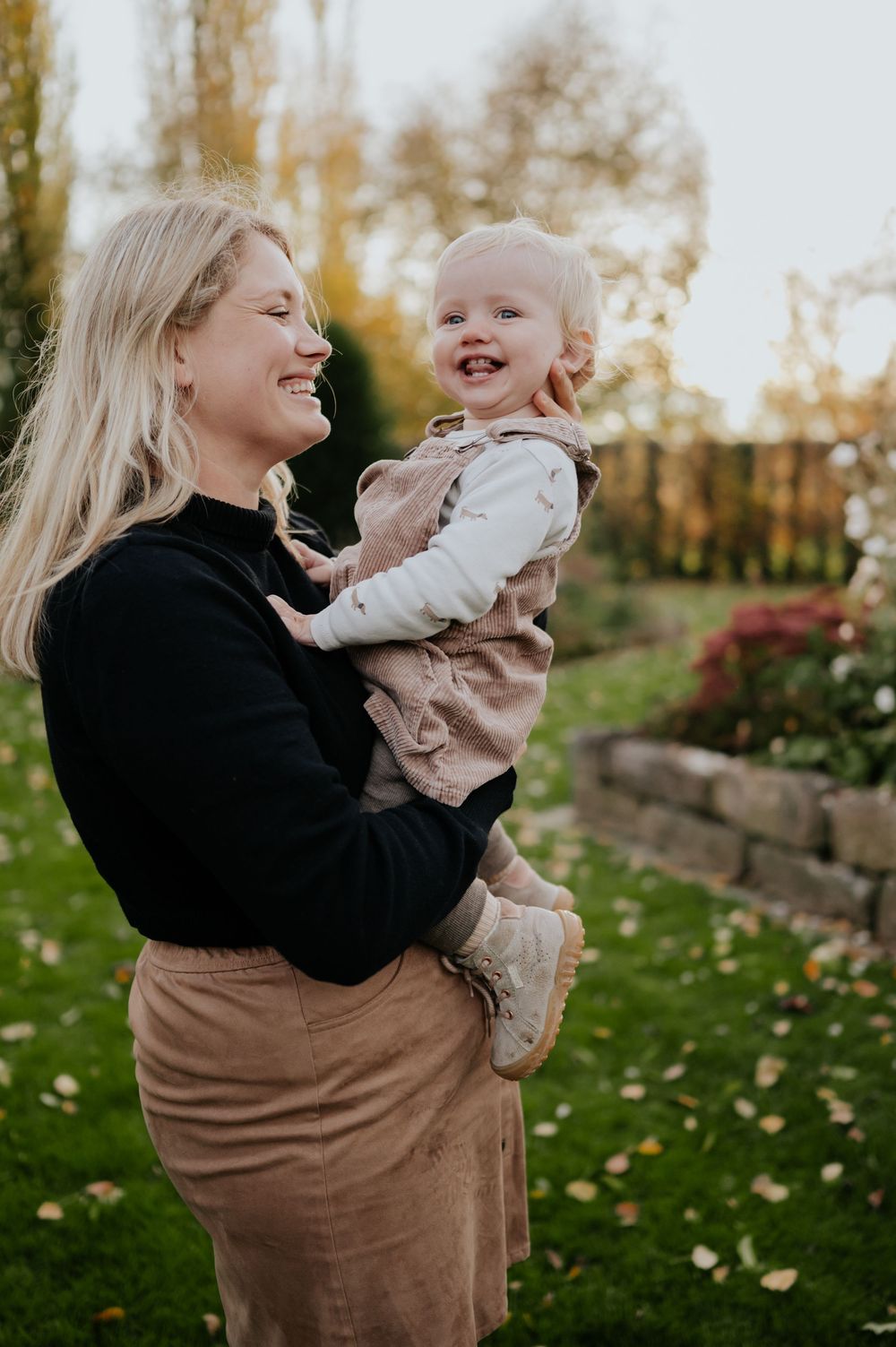 Ein Elternteil in einem schwarzen Pullover und einer braunen Hose hebt ein kleines Kind im Herbst im Freien hoch.