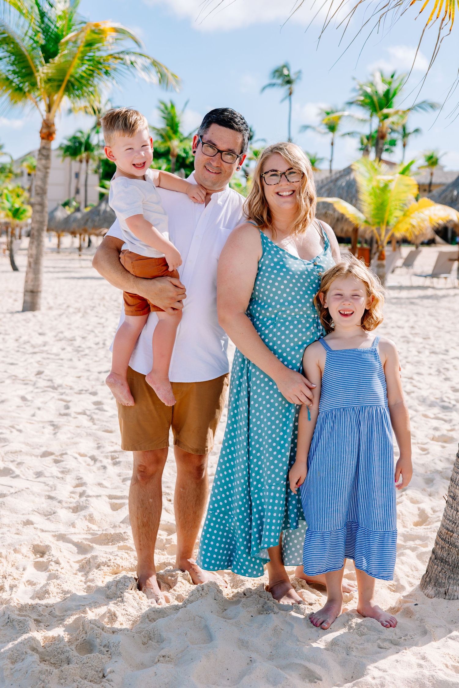 A family wearing coordinated blue and white outfits poses together on a tropical beach with palm trees.