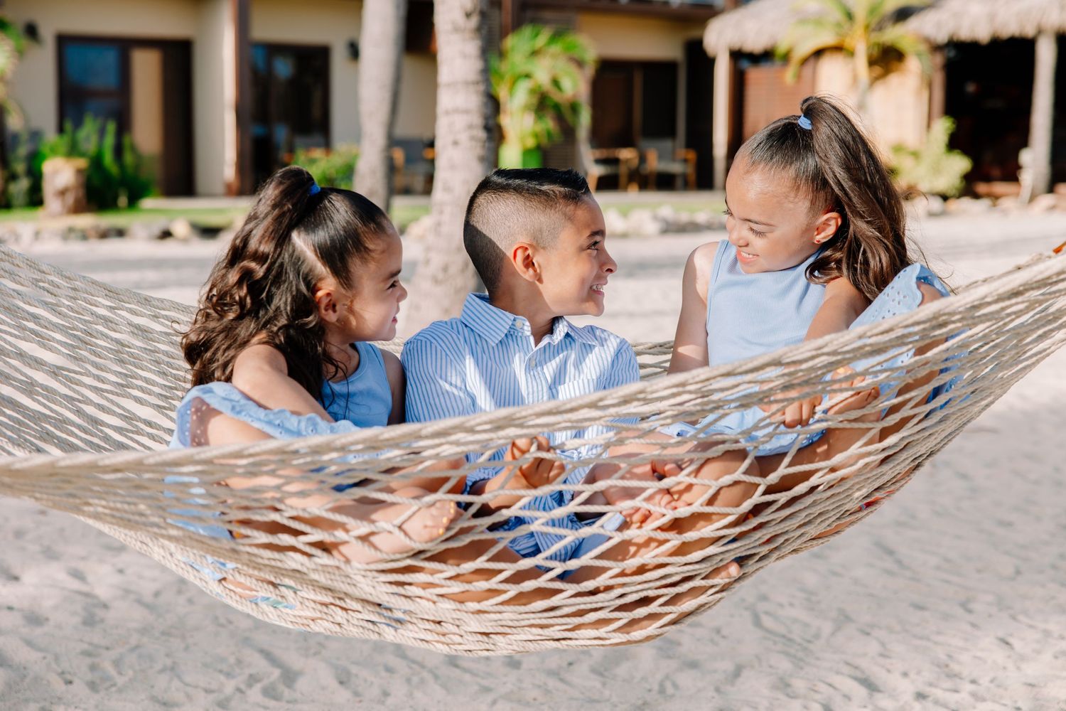 Family relaxing together in a beachside hammock under palm trees.
