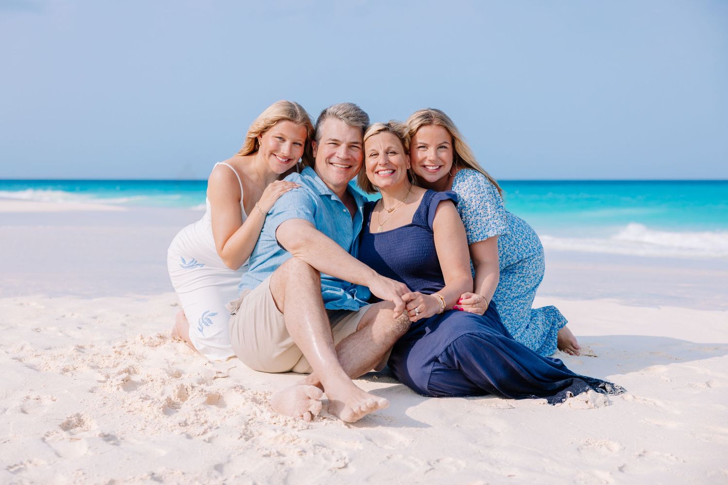 Family group embracing while sitting together on white sand with turquoise ocean backdrop.
