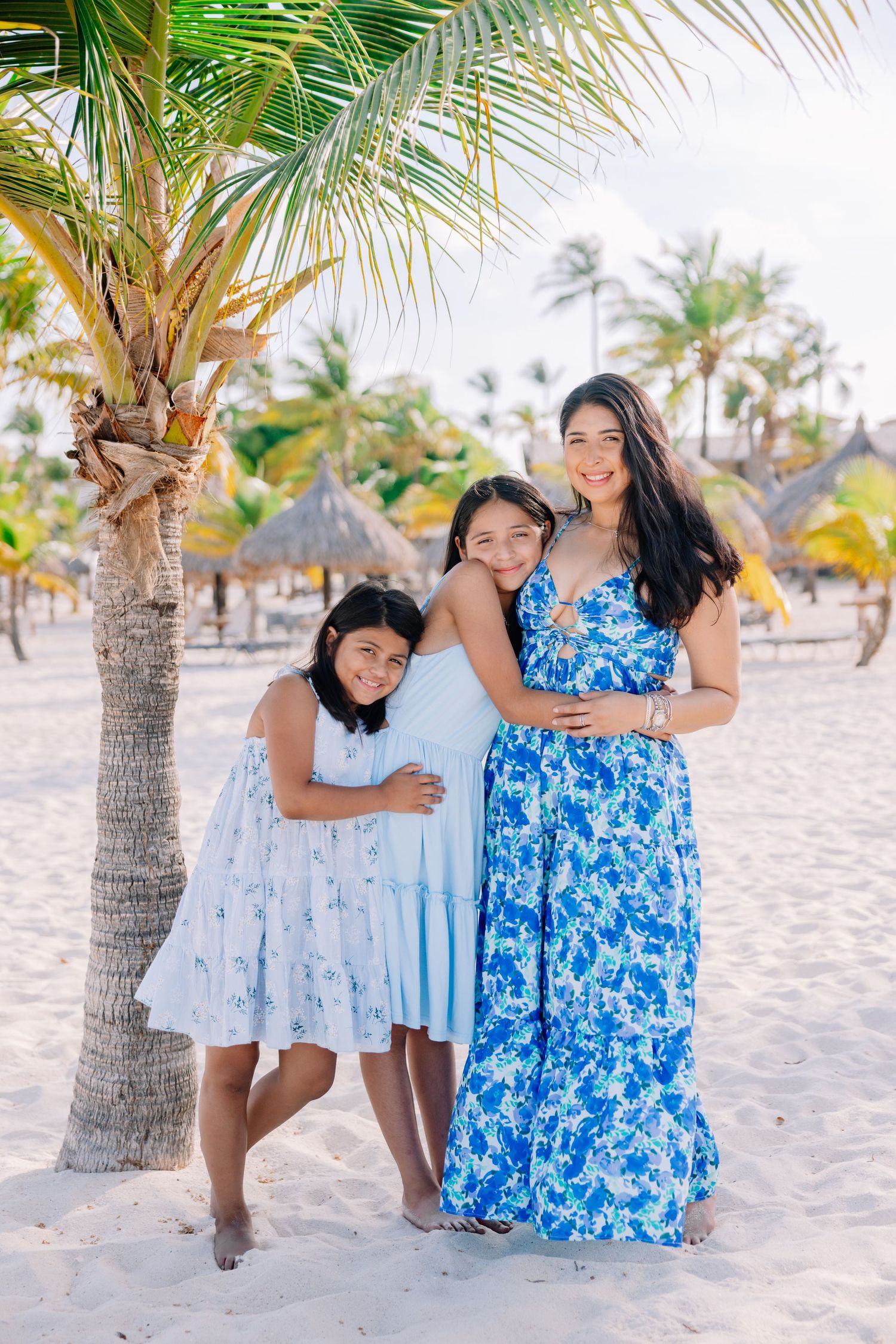 Family members in matching blue and white dresses play together on a sandy beach with palm trees.