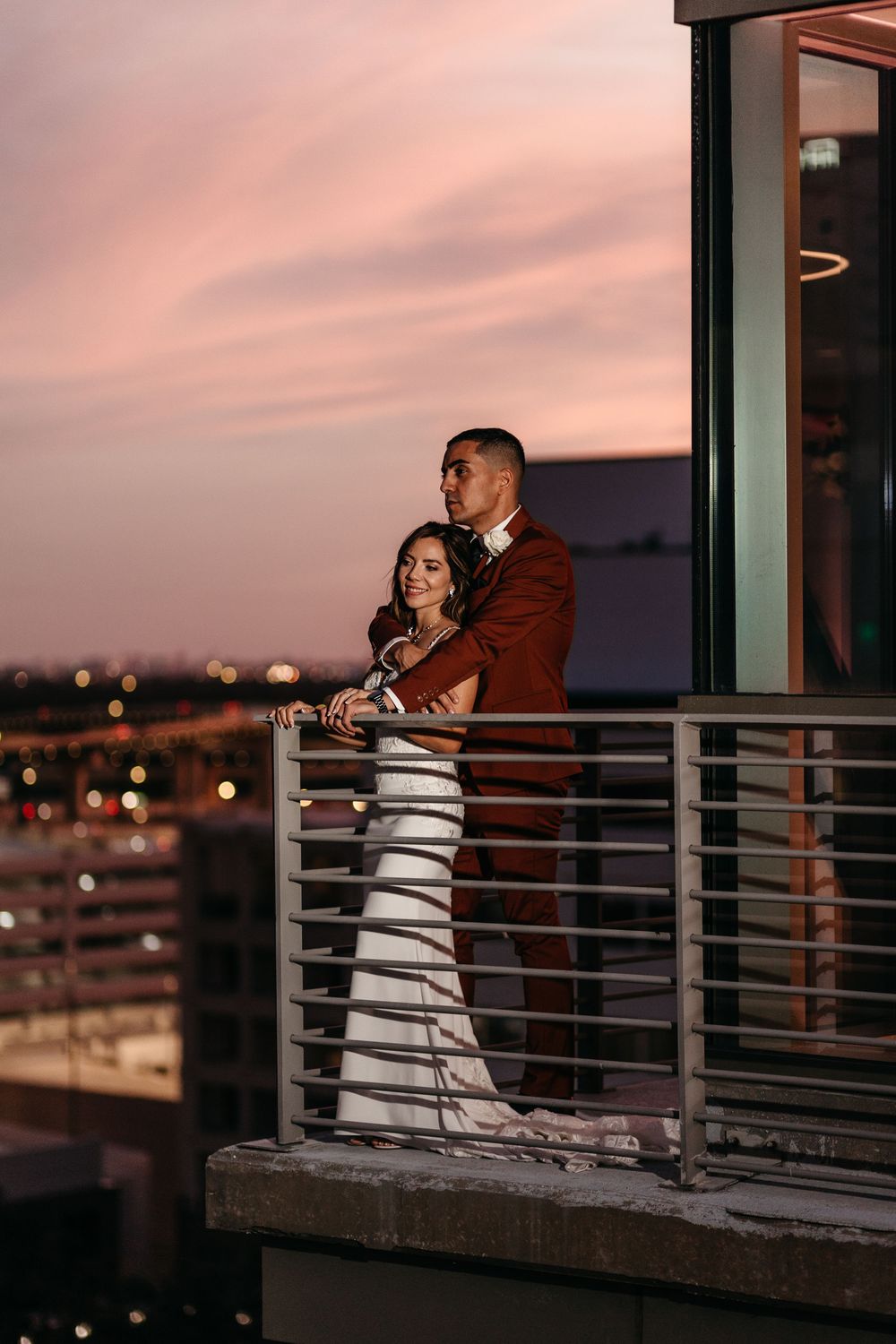 Couple embracing on modern balcony with dramatic pink sunset sky.