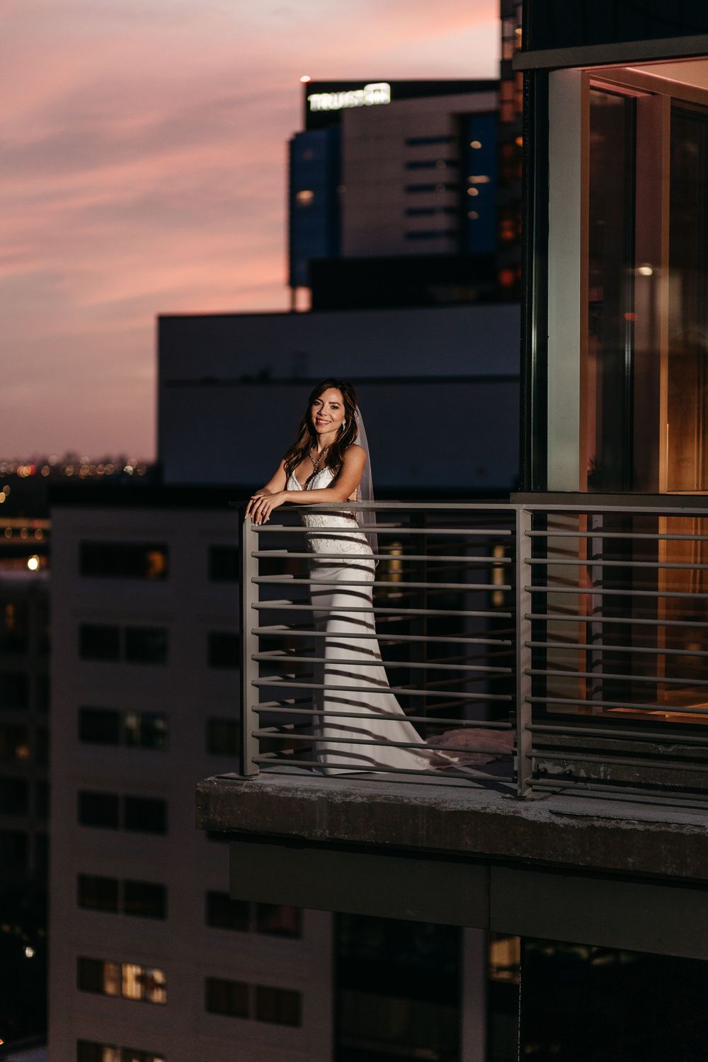 Bride in white dress poses on urban balcony during sunset.