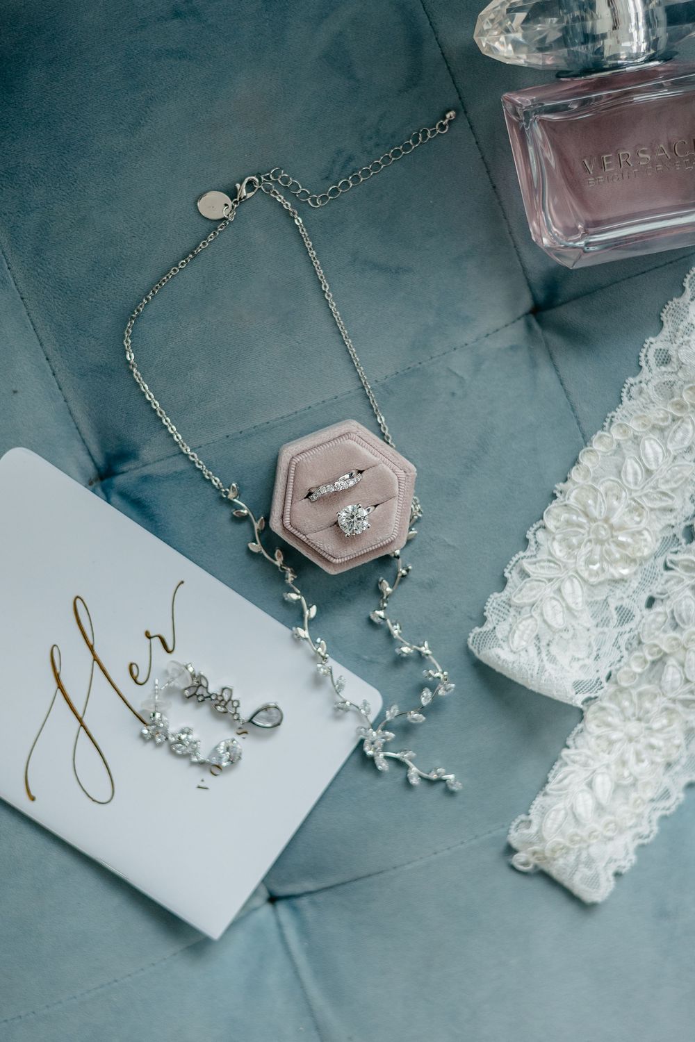 Bridal accessories including jewelry, perfume and vow cards arranged on blue background.