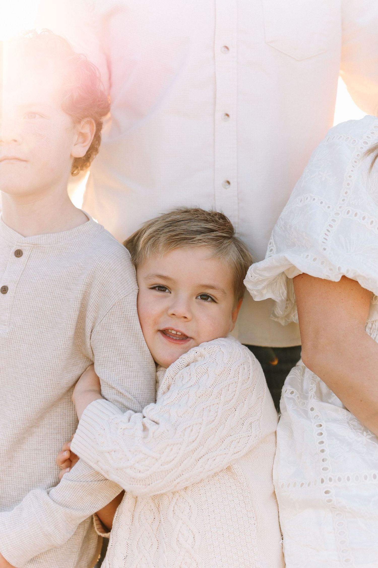 A baby in white lace clothing smiles while holding on to his brother during family photoshoot in California