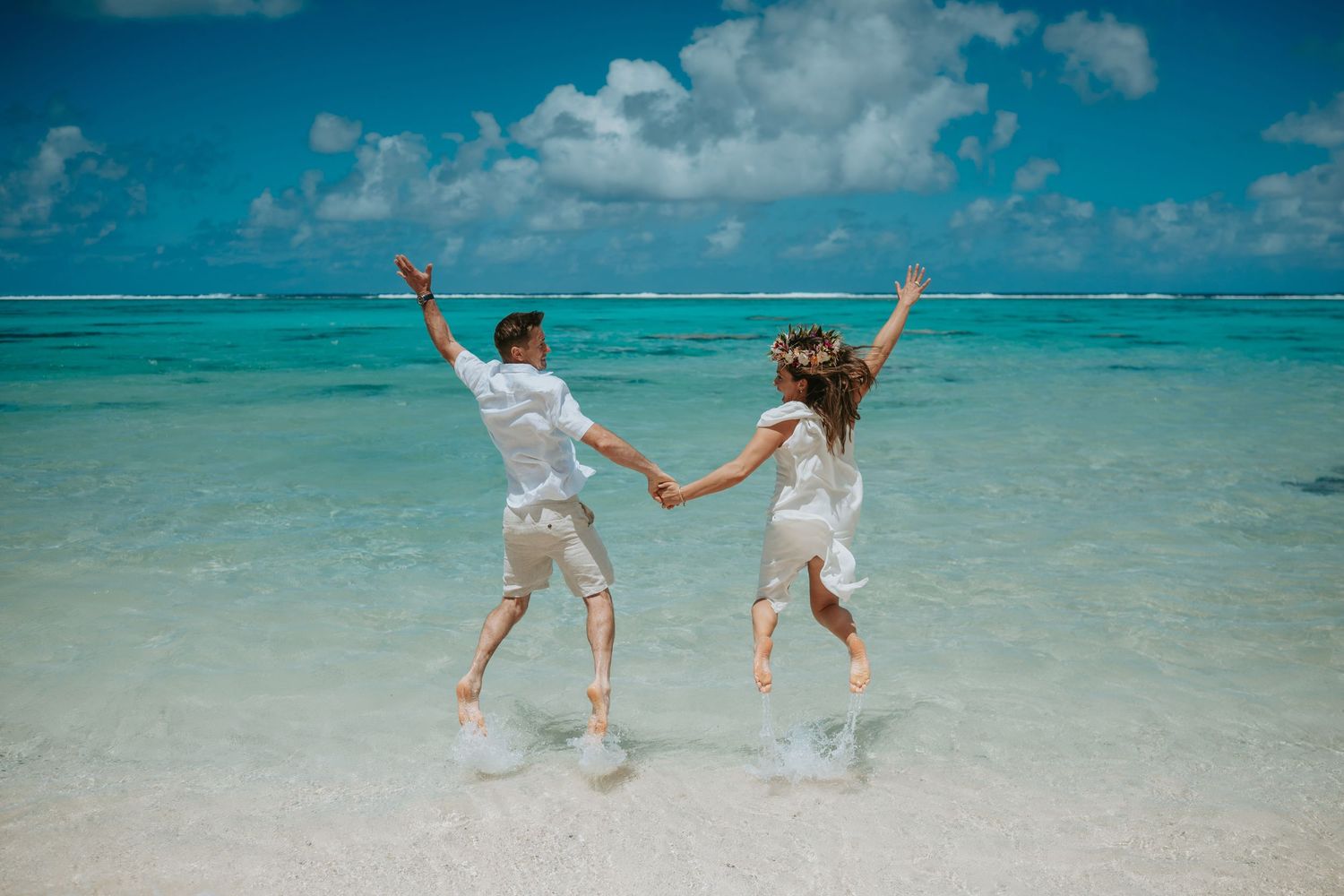 Two people in white outfits jumping joyfully together on a tropical beach with turquoise water and blue sky.
