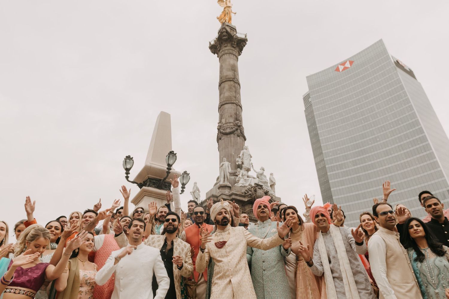 Large crowd celebrating at an outdoor wedding ceremony with city buildings in background.