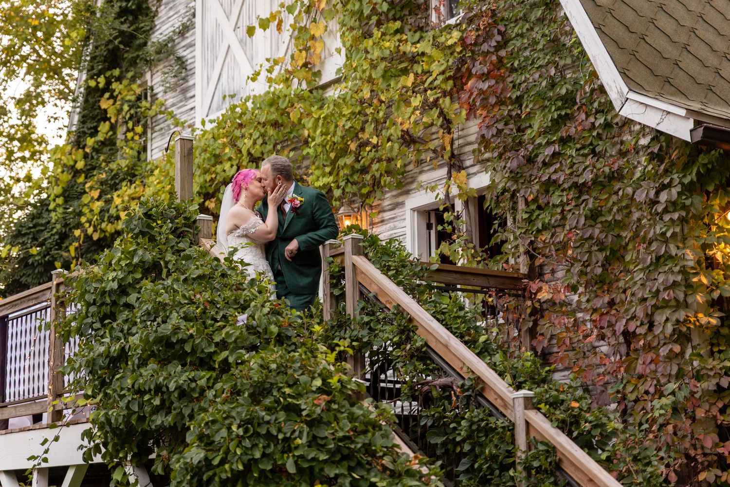 Bride and groom sharing a romantic kiss on ivy-covered stairs outside a charming historic building.