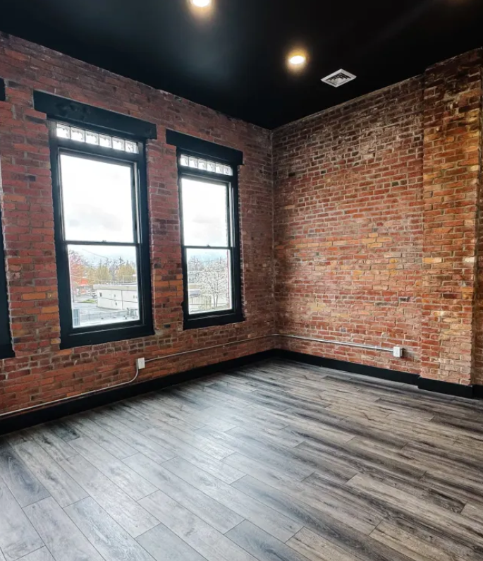 Empty room with exposed brick walls, dark hardwood floors, and two black-framed windows letting in natural light.
