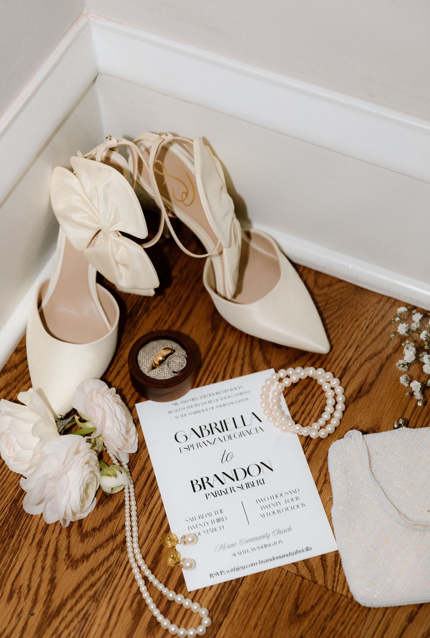 Elegant white bridal accessories including pointed heels, pearl necklace, and wedding invitation on wooden floor.