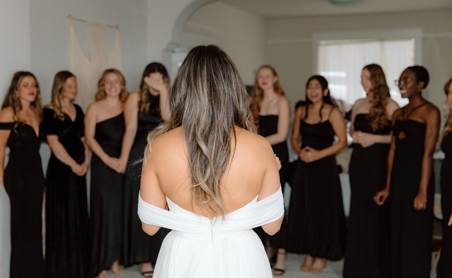 A bride in a white dress stands with her back to a group of bridesmaids wearing black dresses in a bright room.