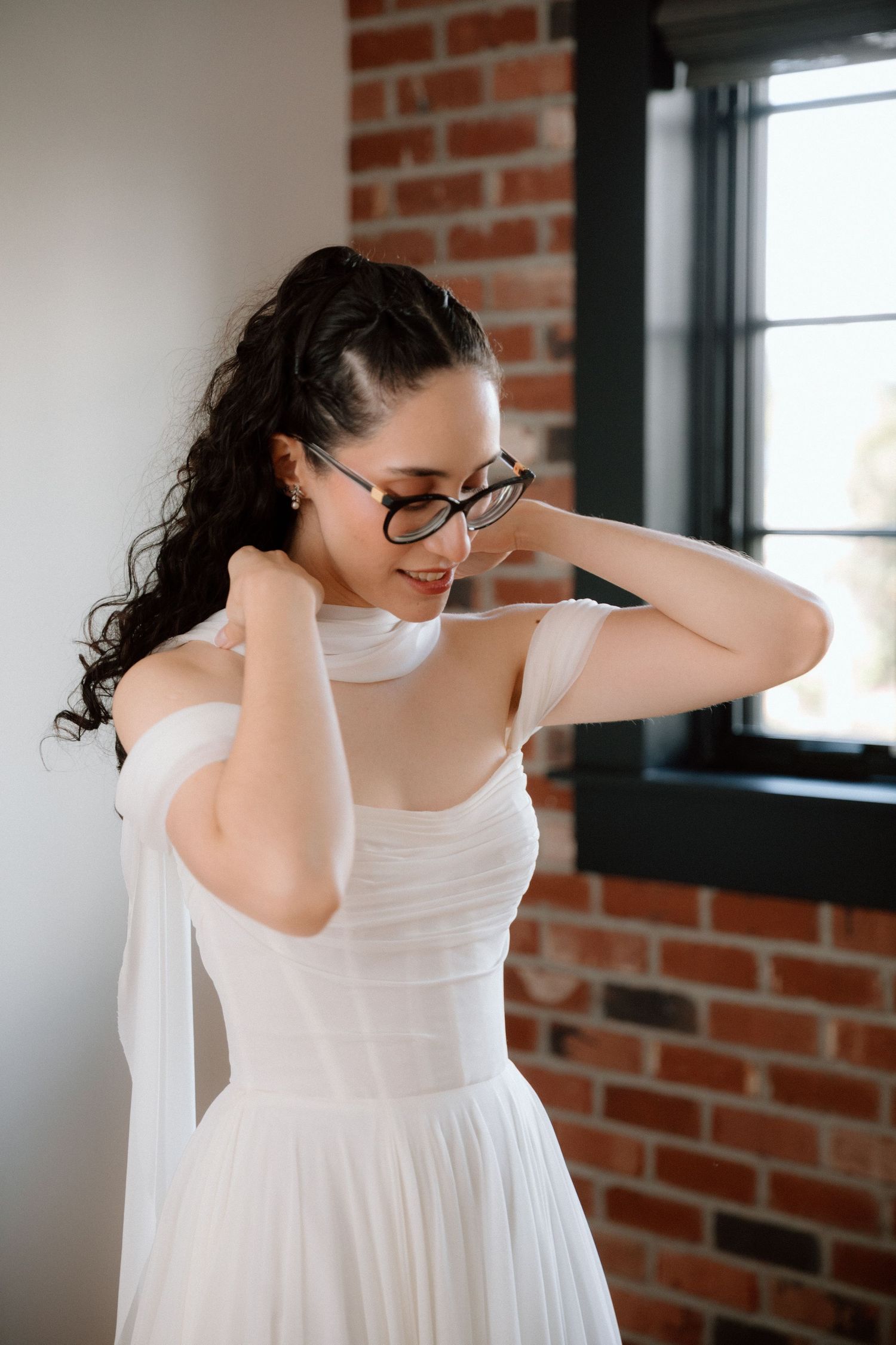 A person in a white wedding dress and glasses stands near a brick wall by a window, touching their neck delicately.