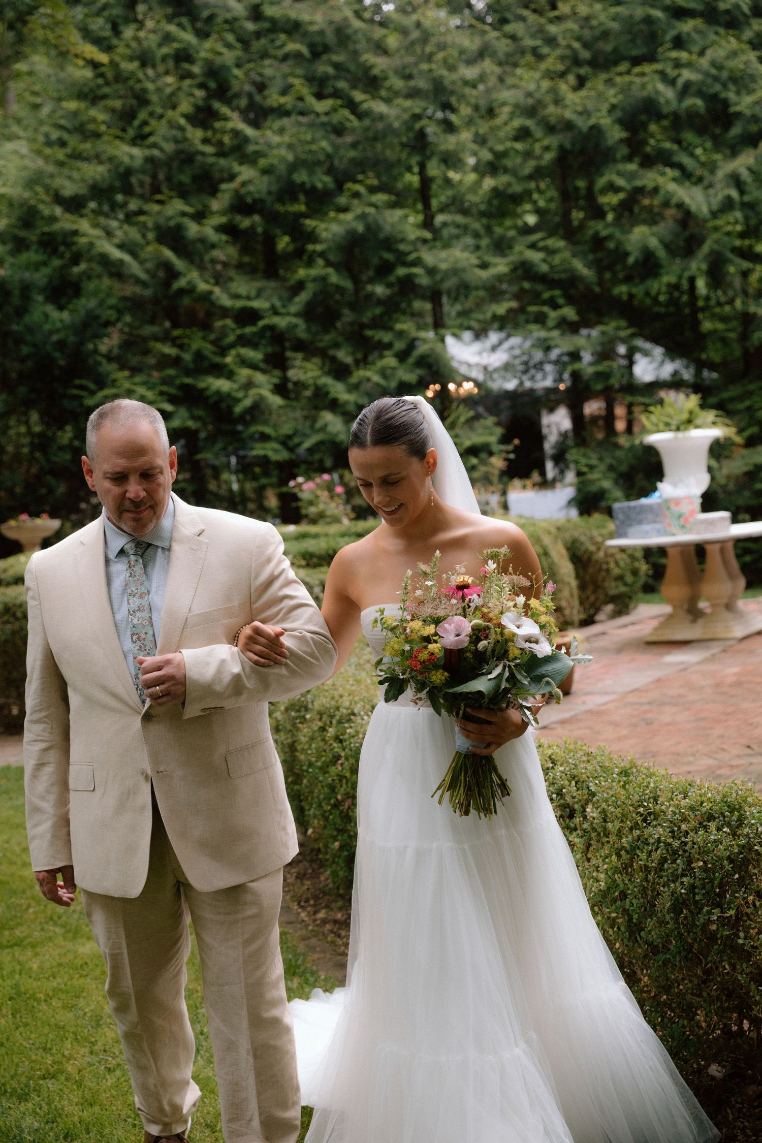 A wedding couple shares a playful moment in a garden setting wearing a white gown and tan suit.