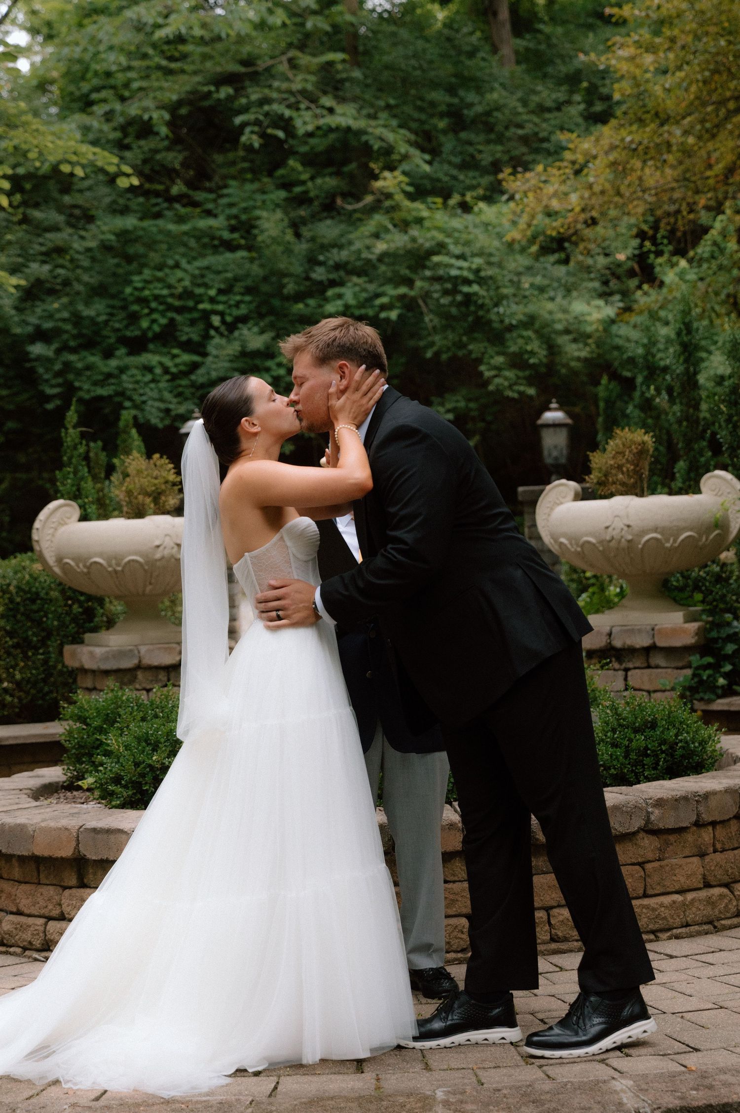 A romantic wedding couple shares intimate moments in a garden setting with stone walls.