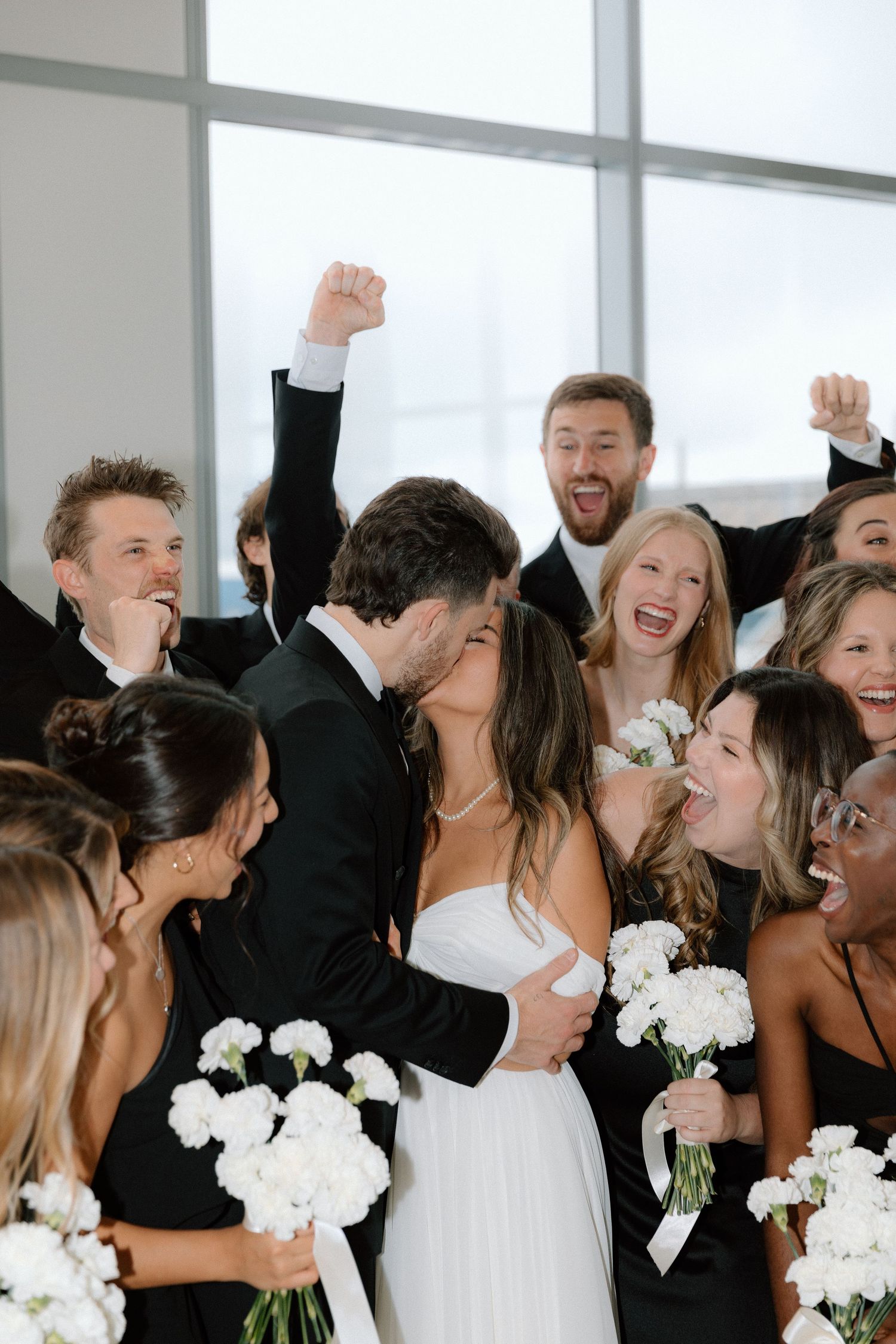 A joyful wedding party in black and white attire celebrates together while holding white floral bouquets.