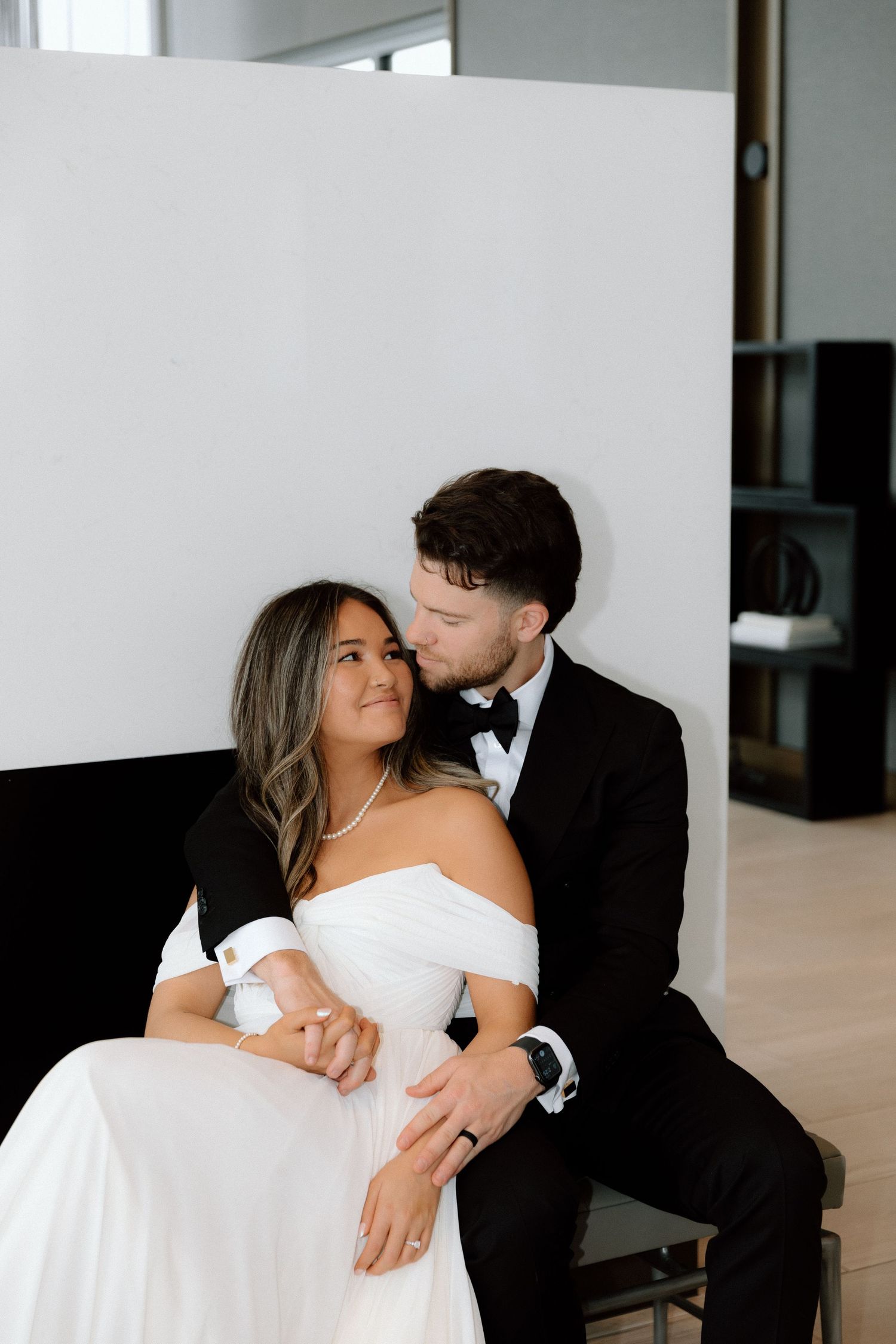 A couple in formal attire share an intimate dance moment against a white wall in a minimalist setting.