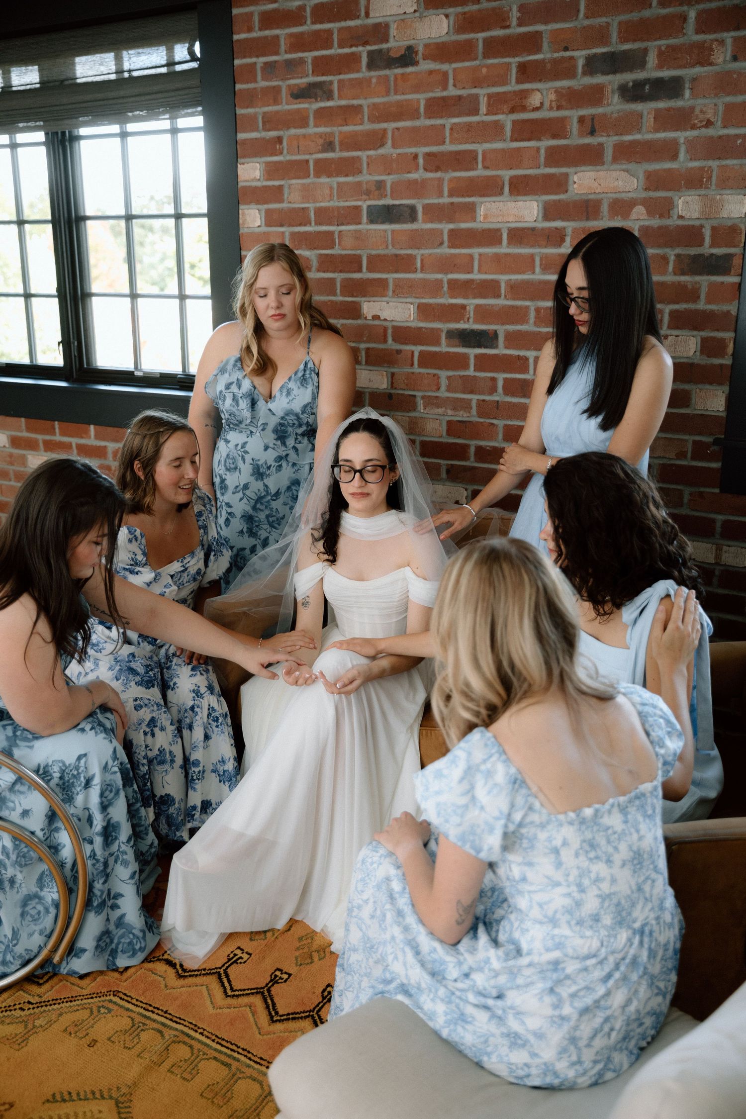 Bridesmaids in blue floral dresses help the bride get ready in a rustic brick-walled room.