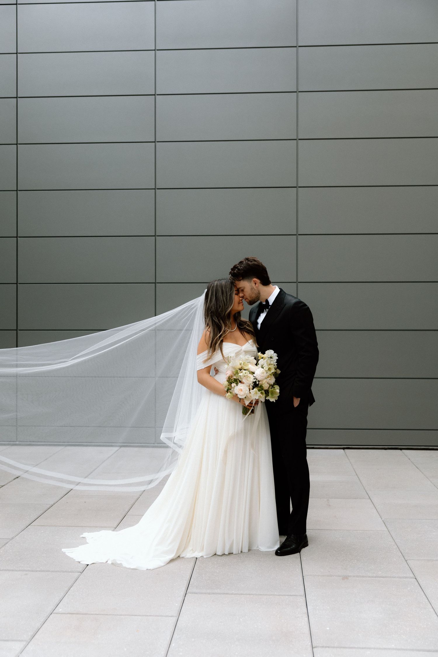 A couple in wedding attire shares an intimate moment as the bride's long veil flows dramatically across a modern grey backdrop.