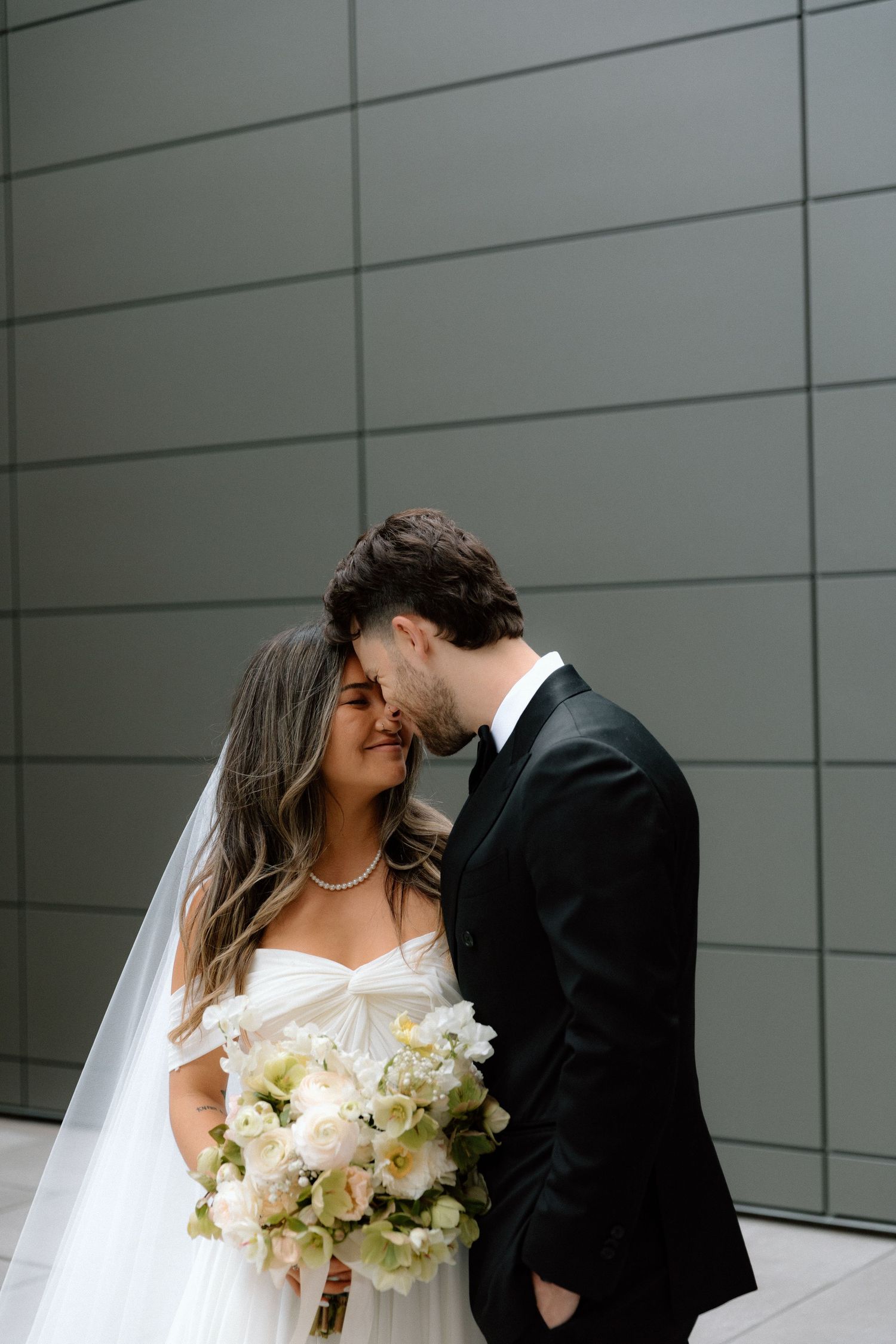 A romantic wedding couple shares a kiss outside a modern building with white bouquet and bridal veil.