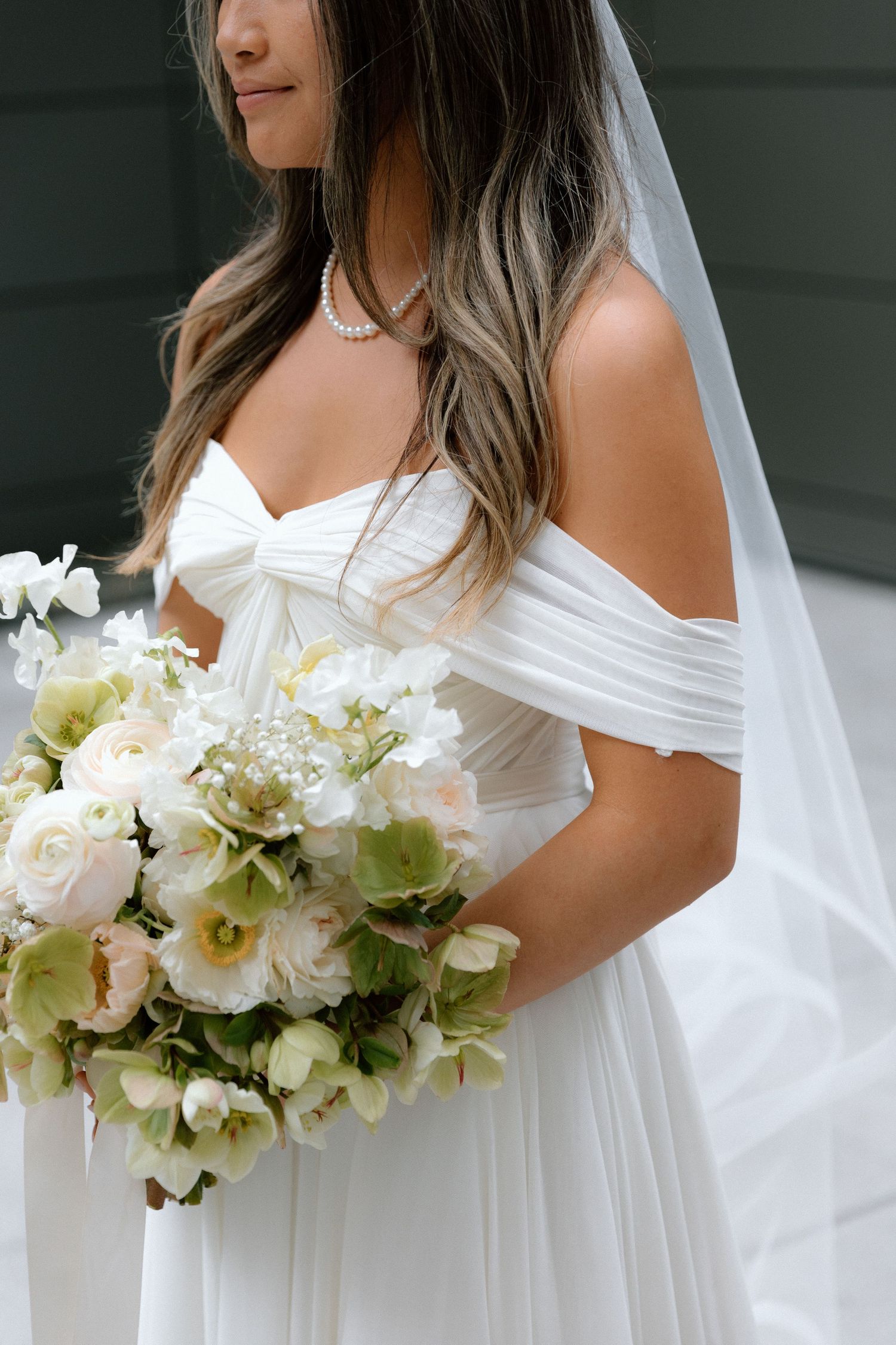 A bridal bouquet of white flowers and greenery is held against a flowing white wedding dress.