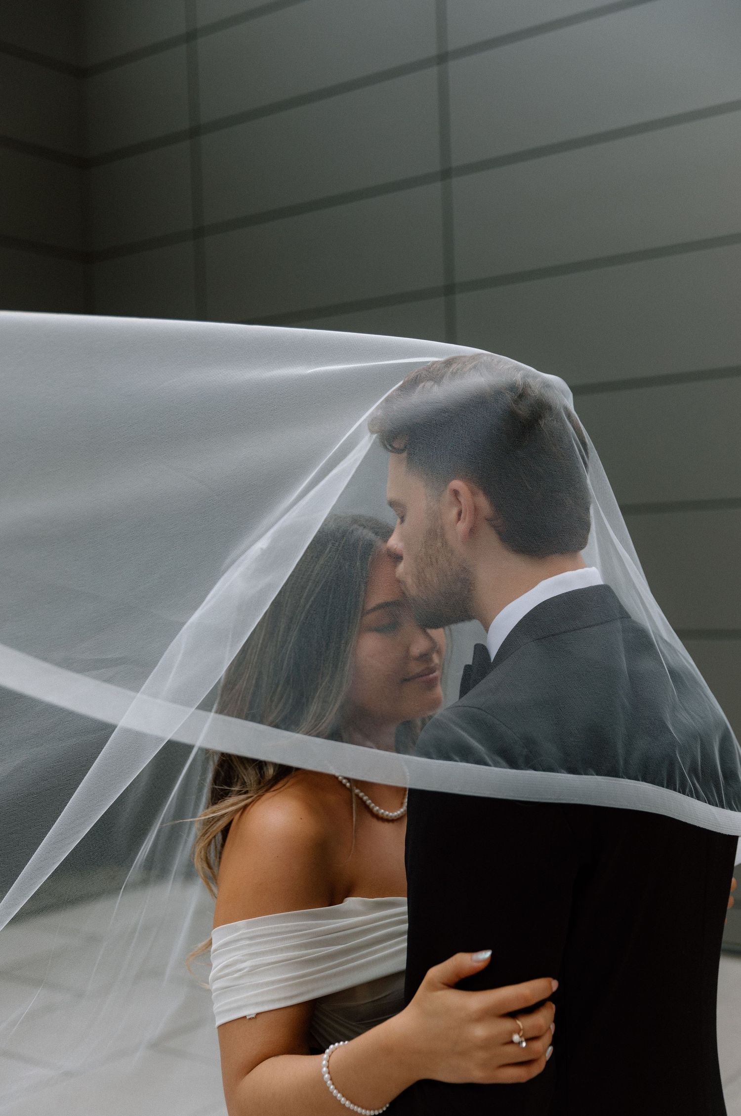 A romantic wedding moment captured under a flowing white veil as newlyweds embrace in an intimate portrait.