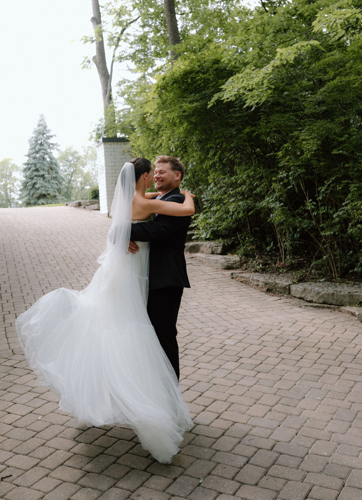 A romantic wedding moment captured on a brick paved path surrounded by green trees and landscaping.
