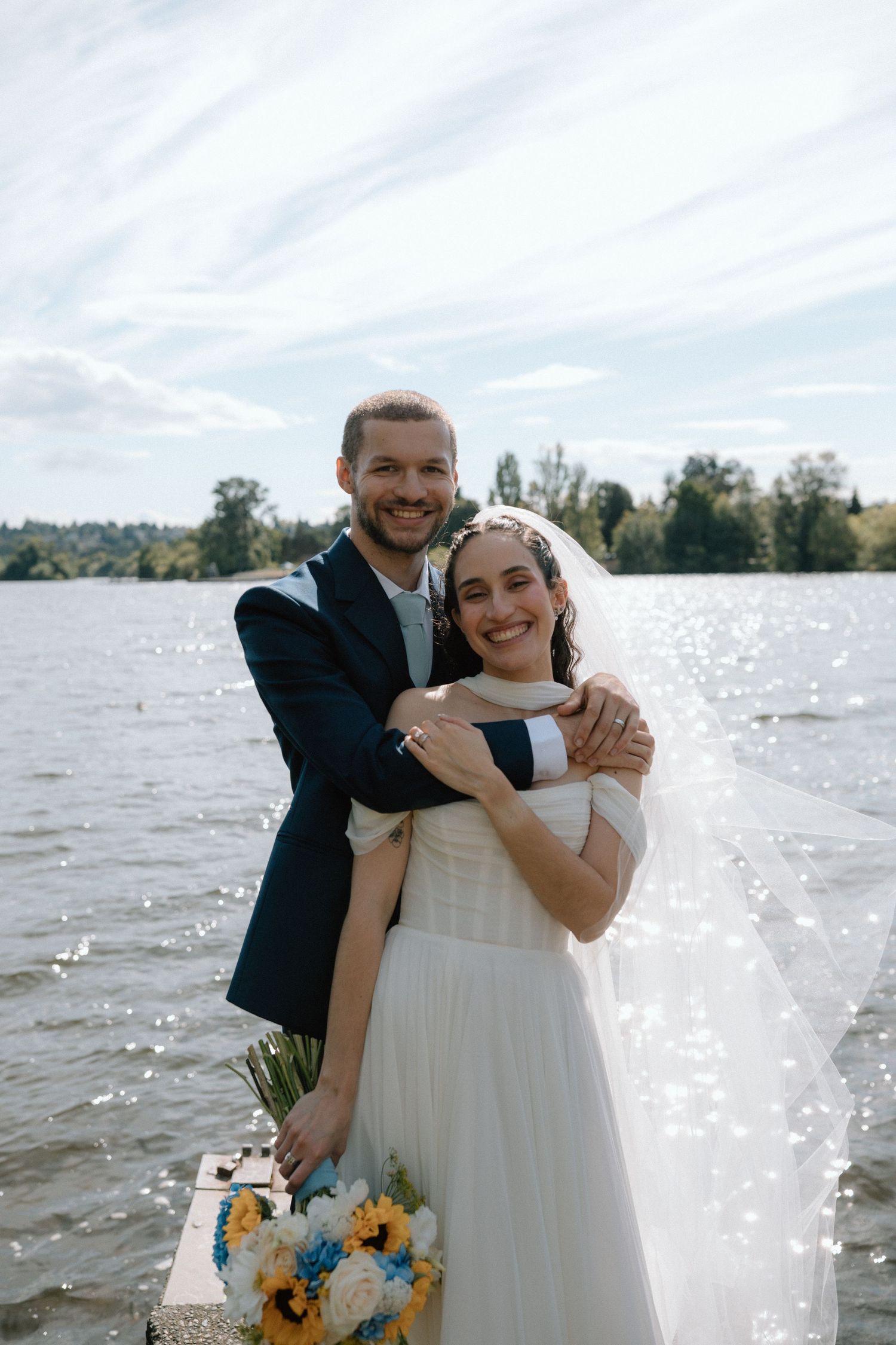 A couple embraces joyfully by a sparkling lake with sunflowers in the foreground on their wedding day.