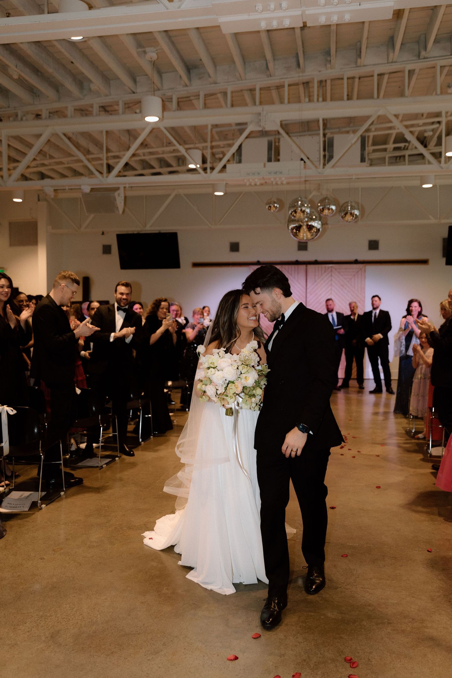 A couple shares their first kiss at a wedding ceremony in an industrial venue with exposed beams and gathered guests.