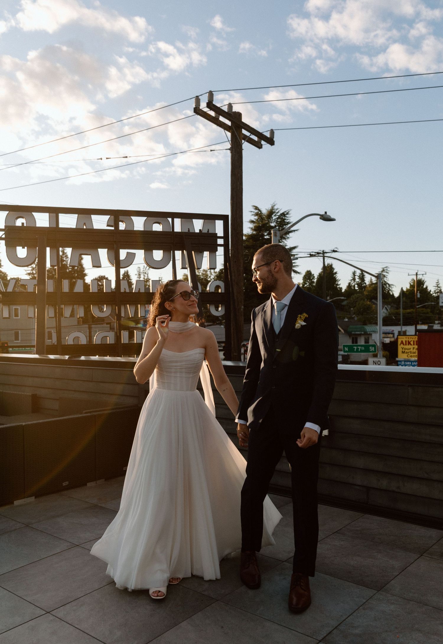 A couple in formal wedding attire stands on a rooftop at sunset with power lines and urban scenery in the background.