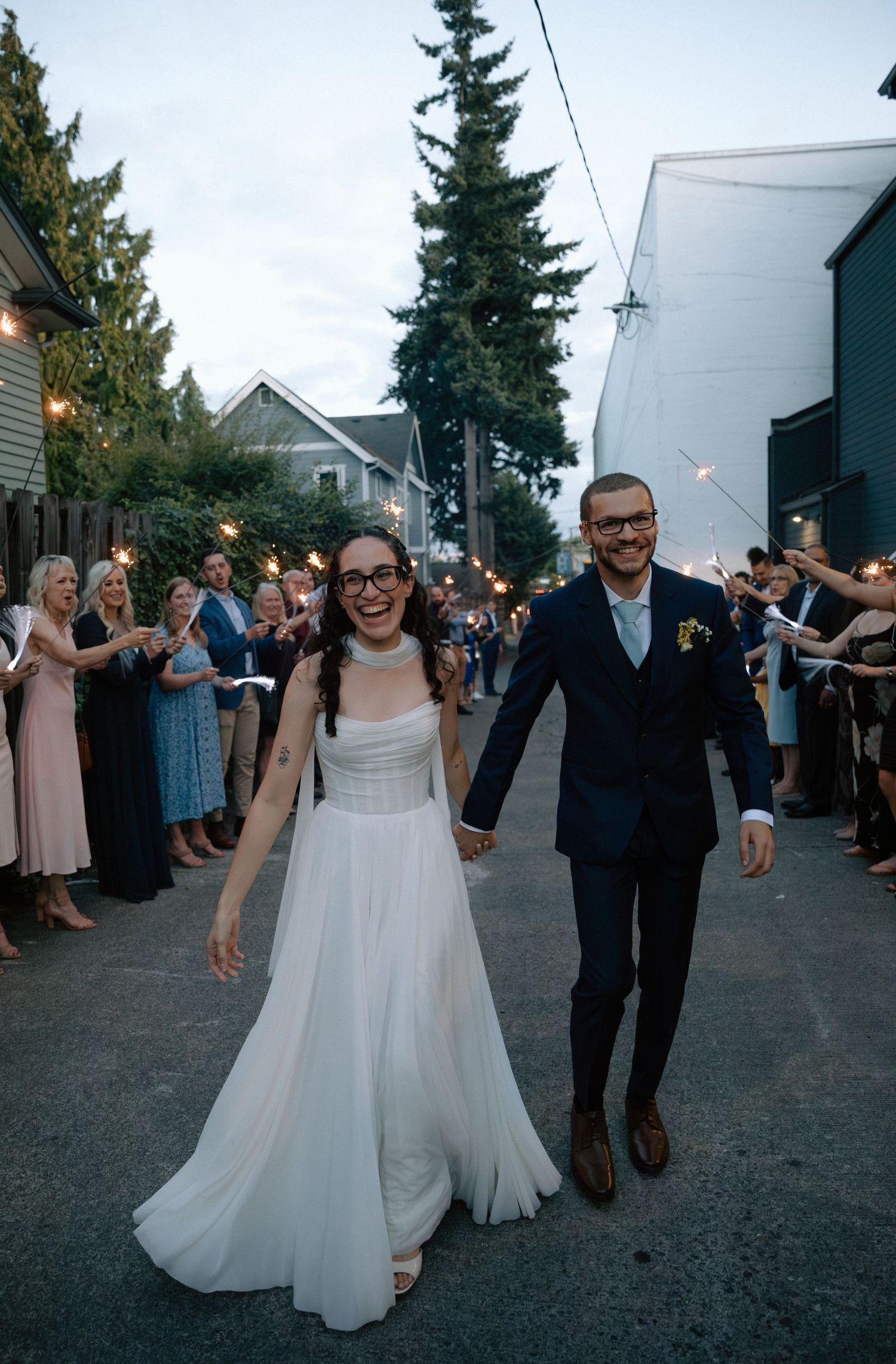 A joyful wedding couple walks hand in hand down an outdoor aisle lined with guests at dusk.