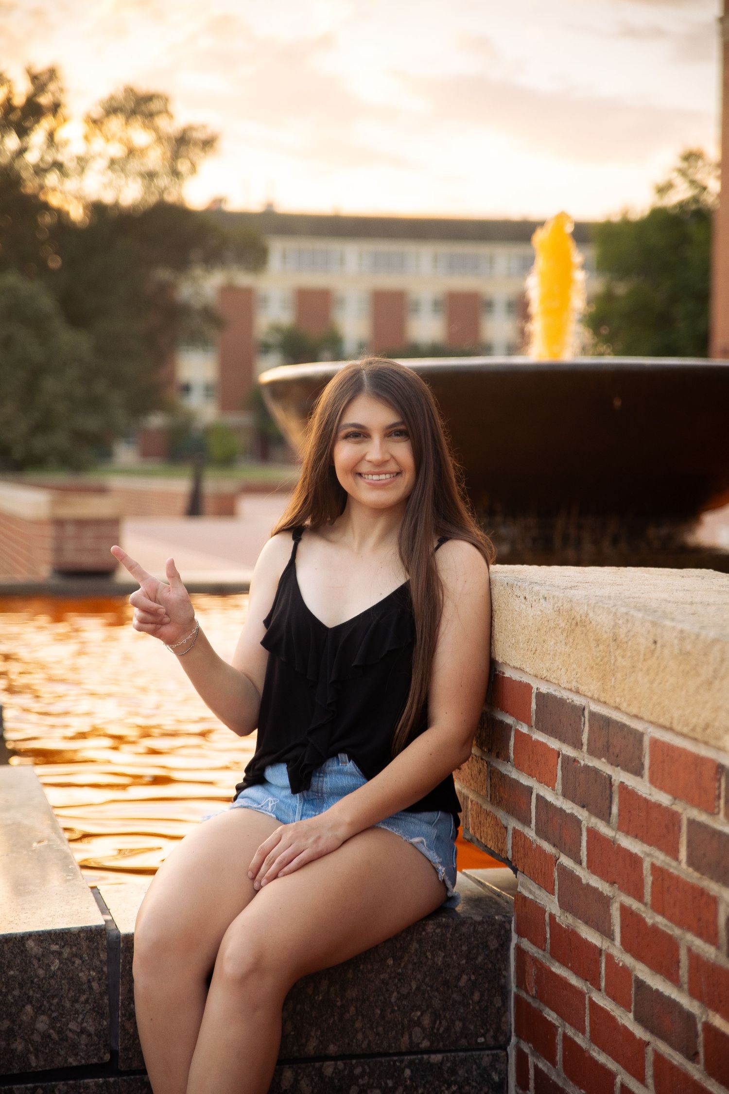 A person in a black top and denim shorts sits on a brick wall during golden hour sunset.